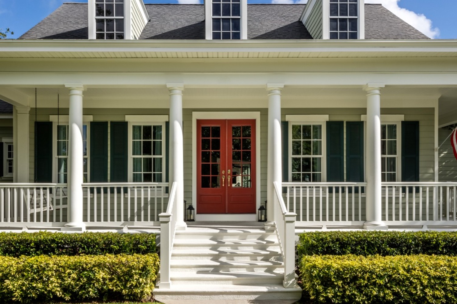 Red double front door on a farmhouse-style home. The siding is gray and there are column supports across the front porch.