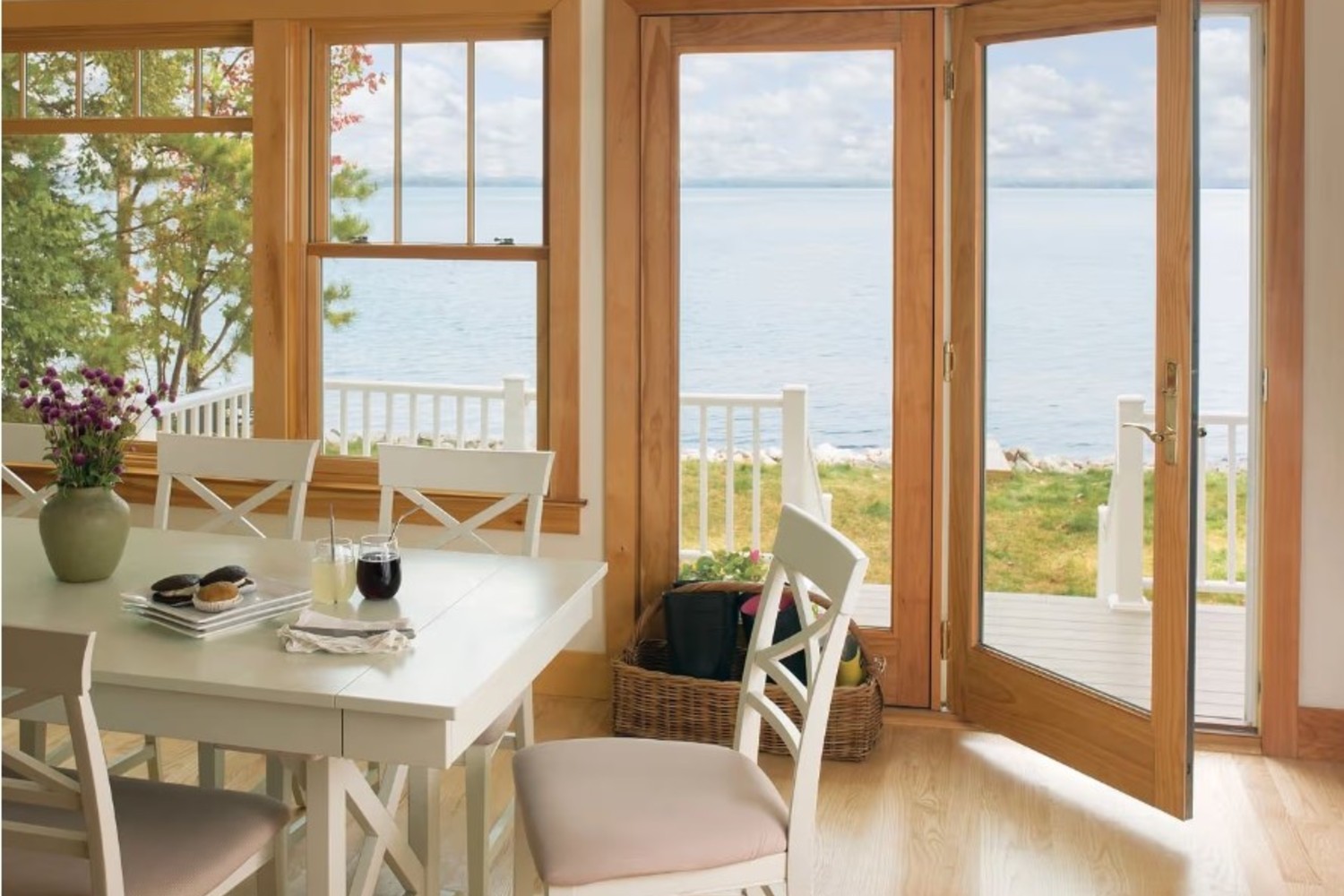 A bright dining area with a white table and chairs sits beside large wooden-framed windows and a glass door overlooking a calm waterfront.