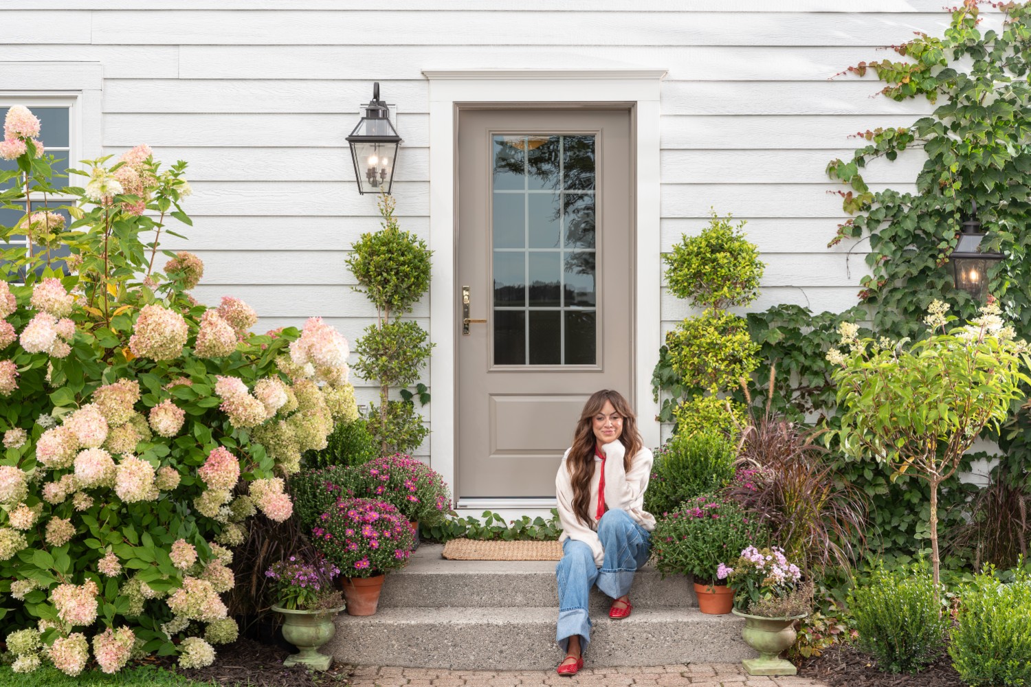 Front entrance of a house with white siding and a beige glass‑paneled door, flanked by black lanterns and surrounded by lush greenery, hydrangeas, potted chrysanthemums, and manicured shrubs. 