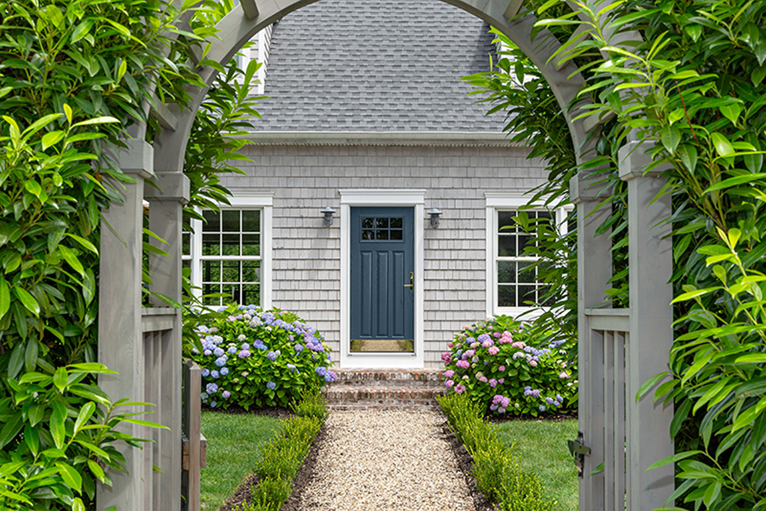 View of a blue front door on a house with gray shakes. The perspective starts under an archway surrounded by tall hedges that follow a short path of paver stones to the front door. 