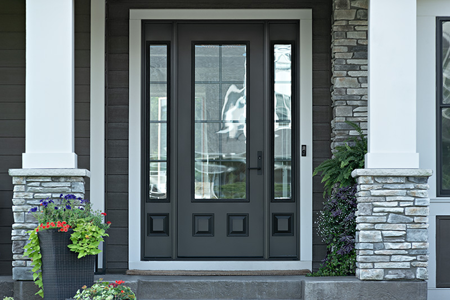 A front doorway to a dark gray house with a black front door framed by white trim. There are two white columns with stone bases in front of the doorway.