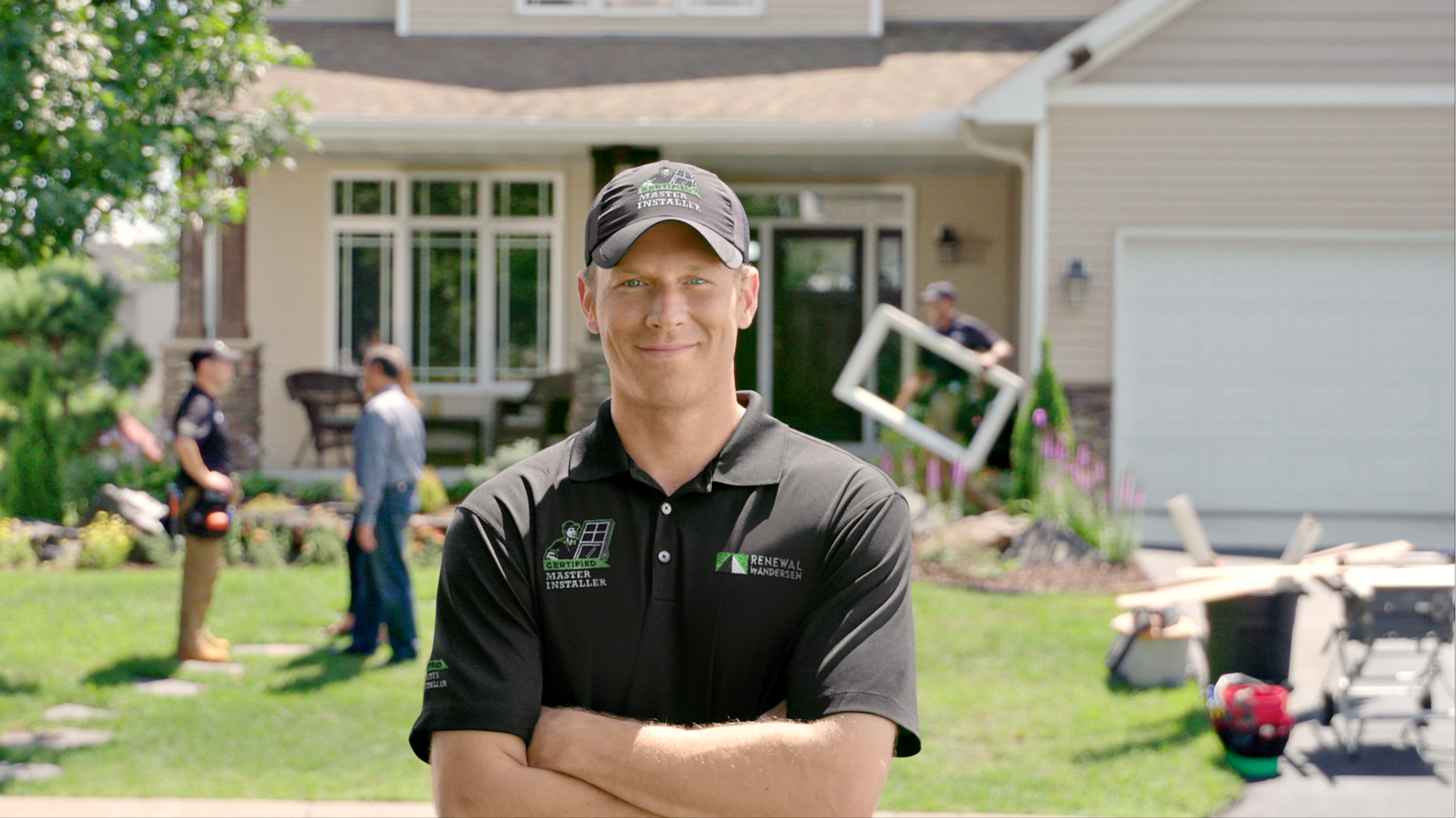 A man wearing a black shirt and hat stands in front of a house, smiling at the camera.