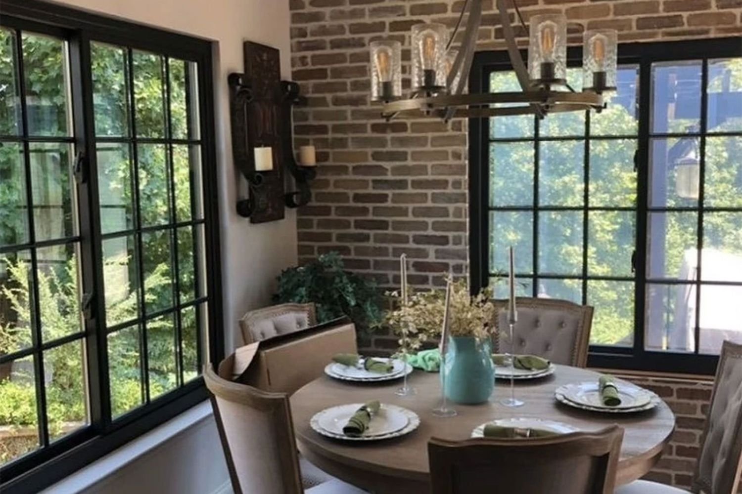 Dining room table set for 5 in the corner of a room with a brick wall and a tan wall with black sliding windows on either wall.