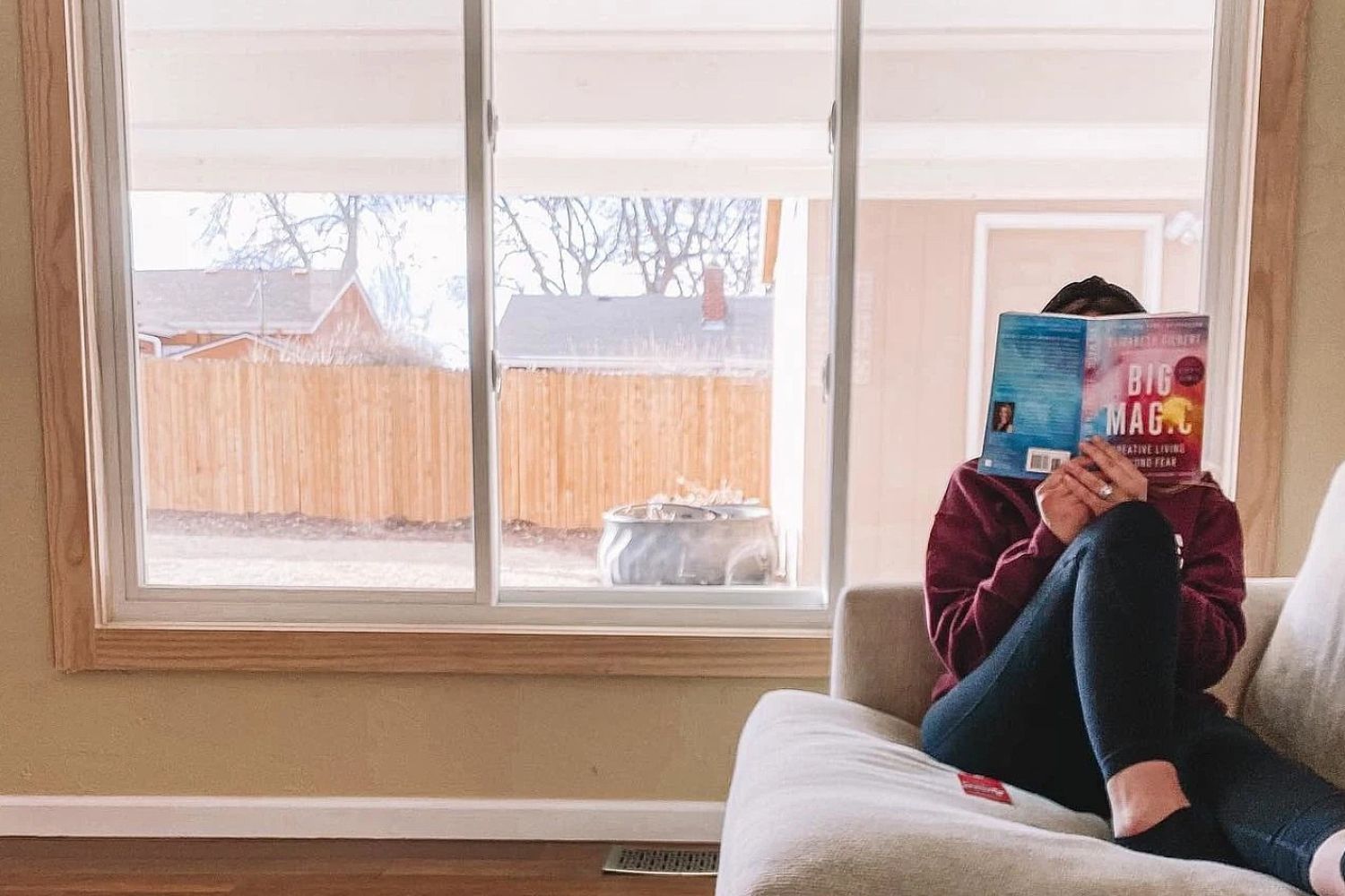 A woman sitting on a couch reading a book. Behind her is a white sliding window.