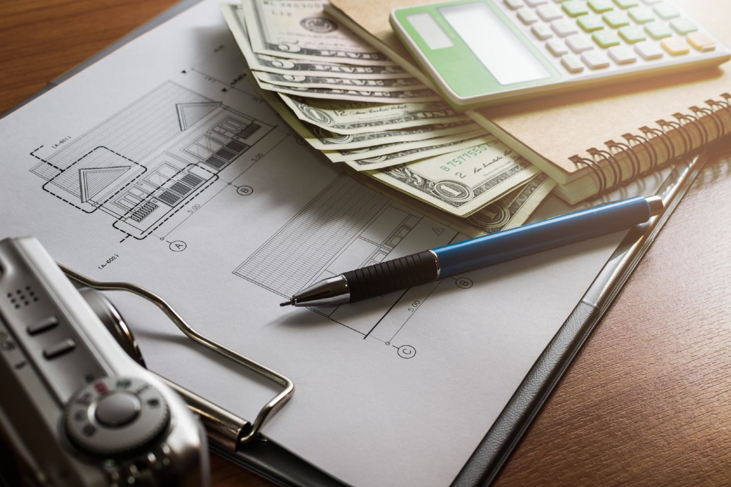 Home design plans on a clipboard with a notebook, a calculator, a pen, and money on top of it all sitting on a wooden table. 