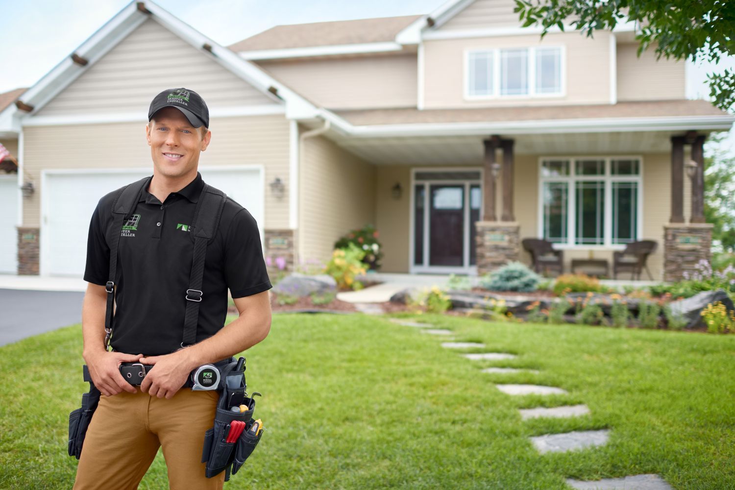 A man wearing a black shirt and pants stands in front of a house, smiling and looking towards the camera.