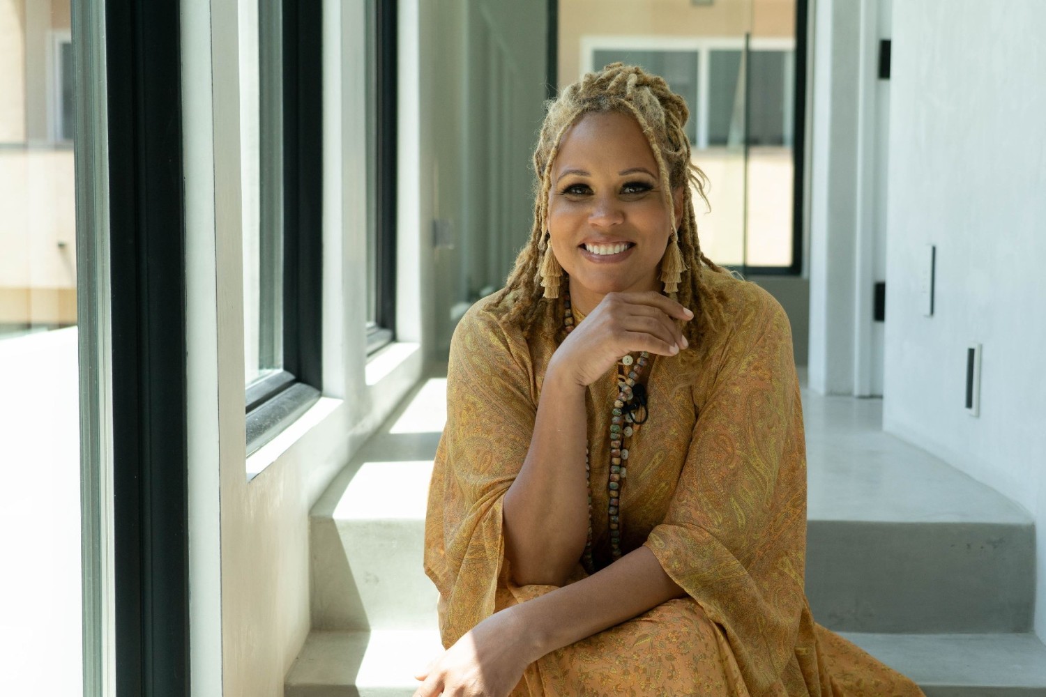 Breegan Jane sits in the hallway of her beach house. She is flanked by several large picture windows with black frames.