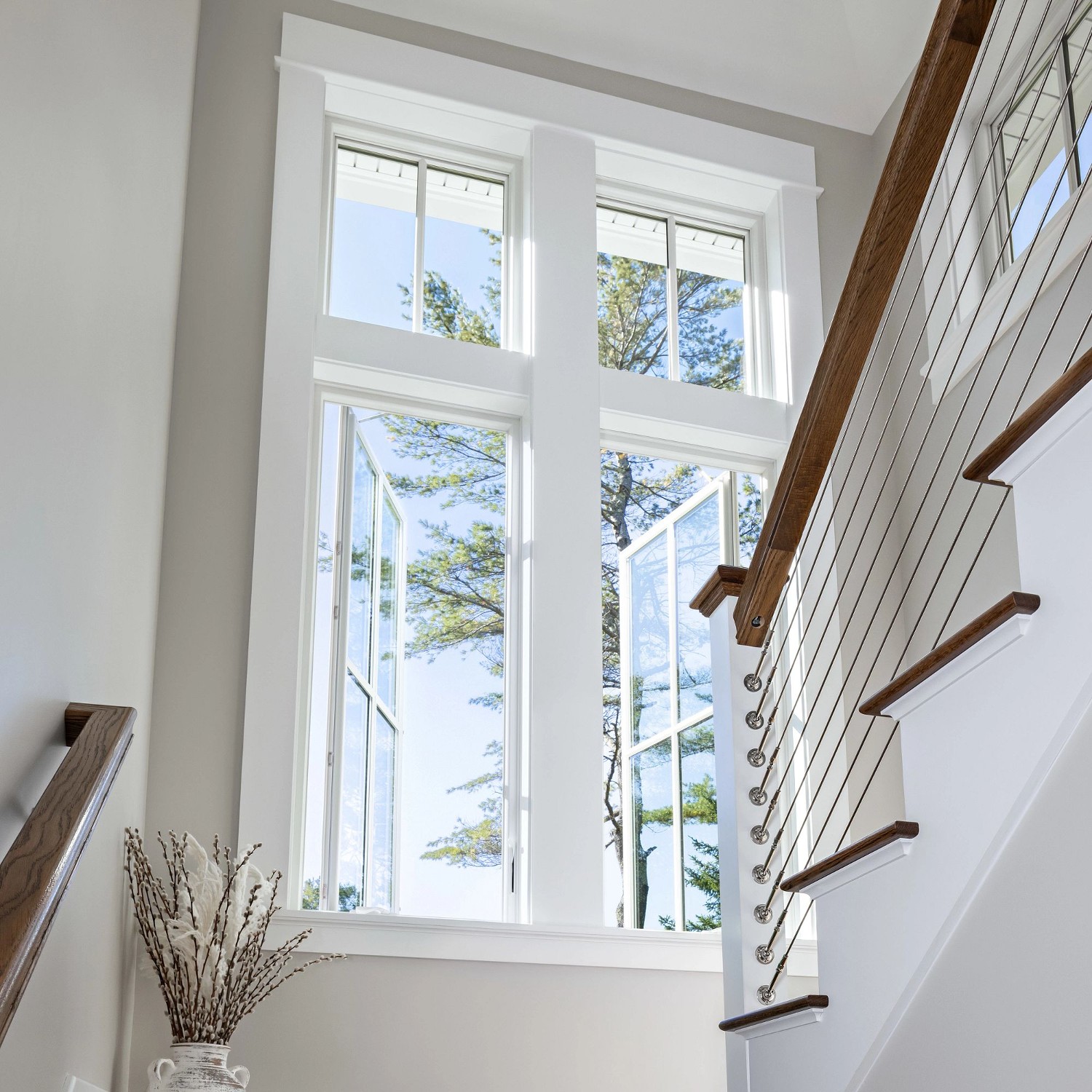 Looking upward into a stairwell with a set of large white casement windows topped with picture windows with grilles.